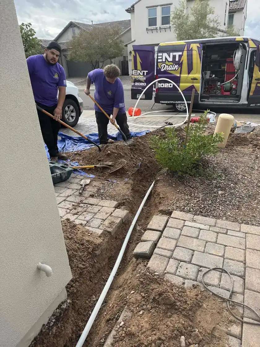 A plumber excavating and inspecting a main water line in a trench next to a brick walkway.