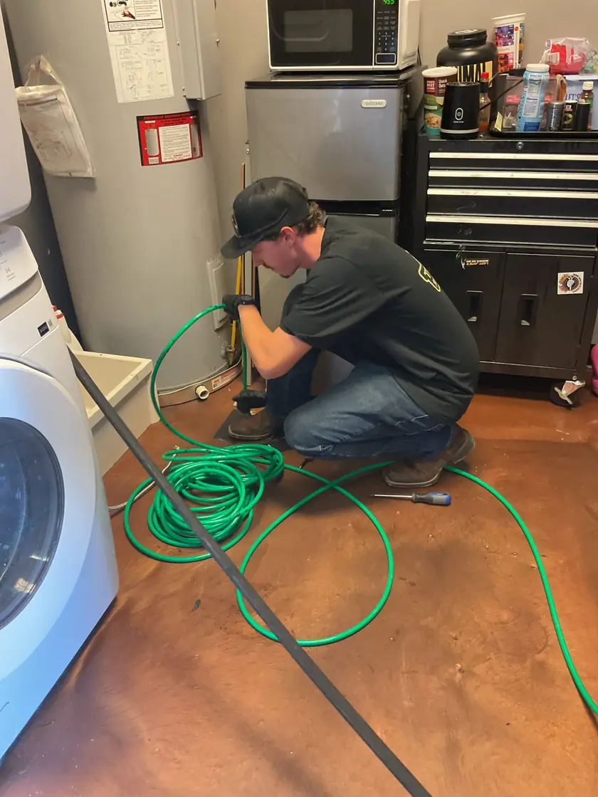 A plumber kneeling on a tile floor while carefully feeding a flexible green hydro jetting hose into a drain pipe cleanout.