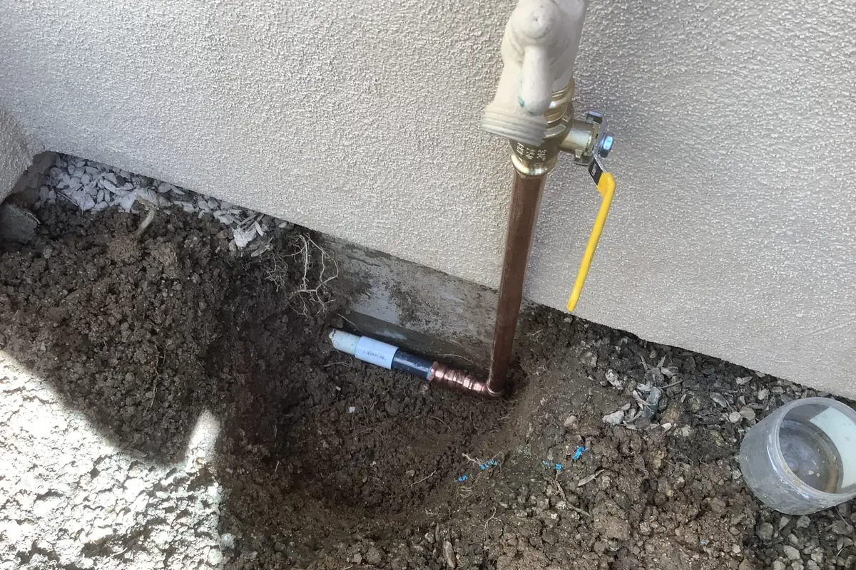 A main water line rising from the soil to a shut-off ball valve with a yellow lever at a stucco wall