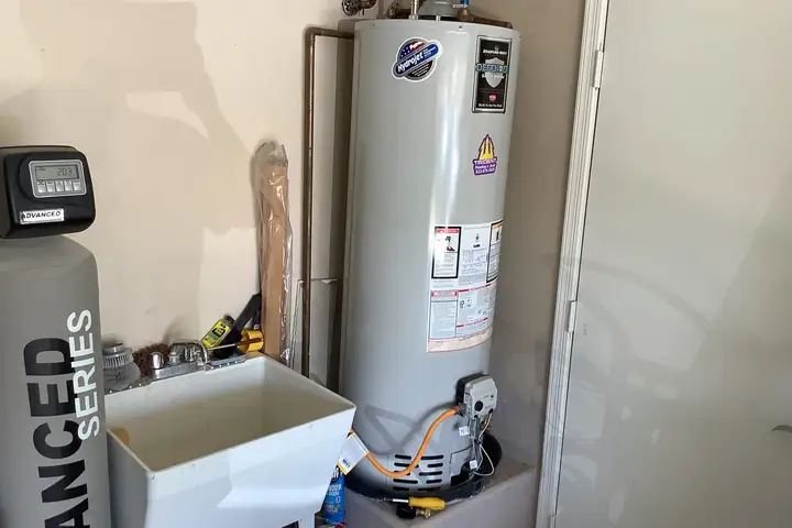 A gas tank water heater on a raised platform in a garage, next to a water softener and a utility sink