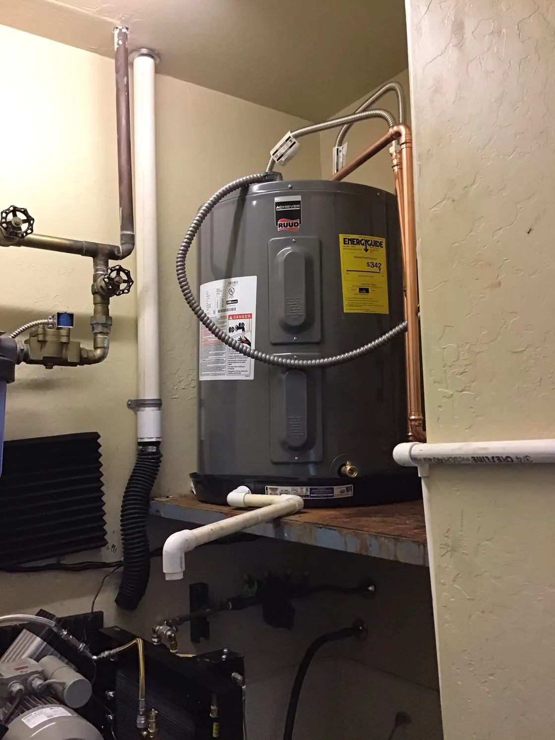 An electric tank water heater installed on a shelf in a mechanical space with copper and PVC piping
