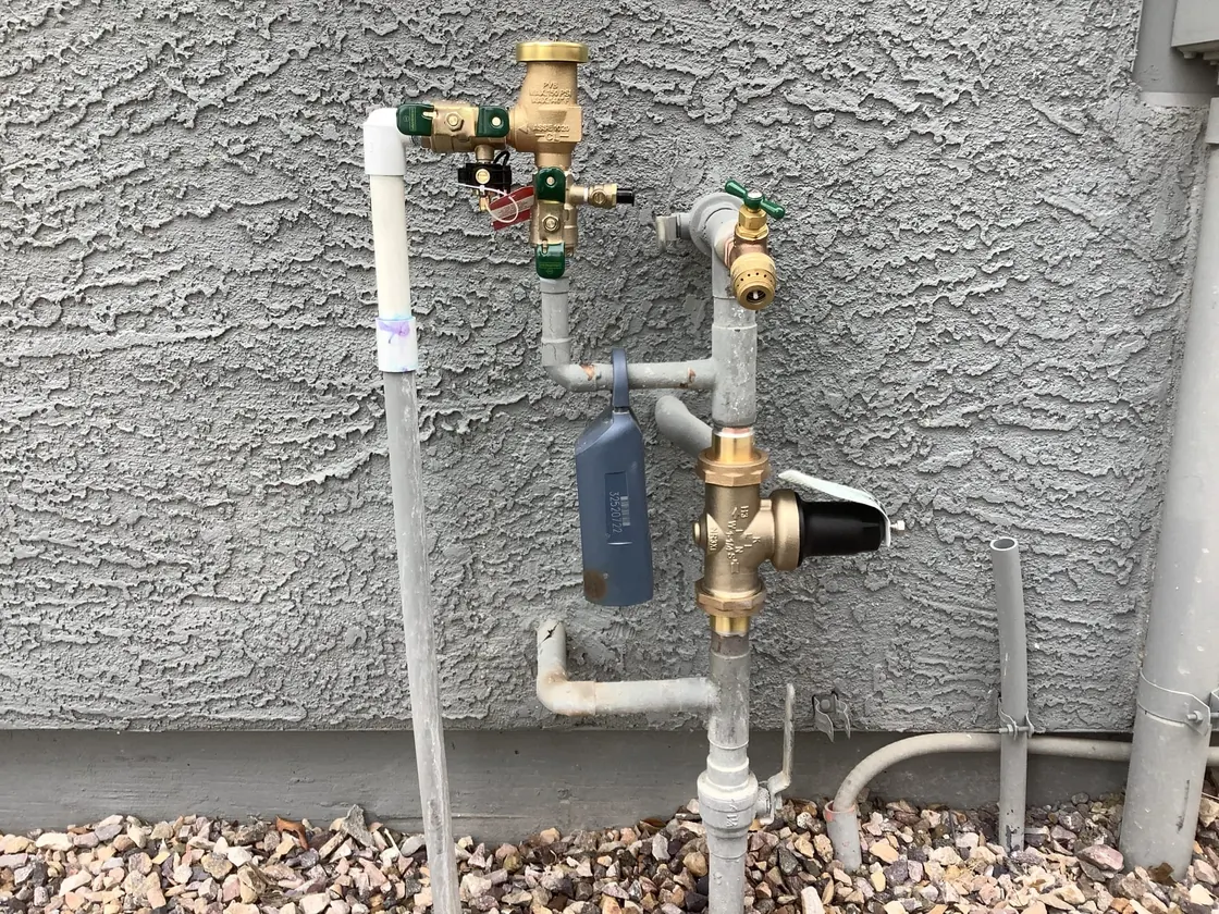Exterior water supply piping with a pressure regulator and valves on a gray stucco wall