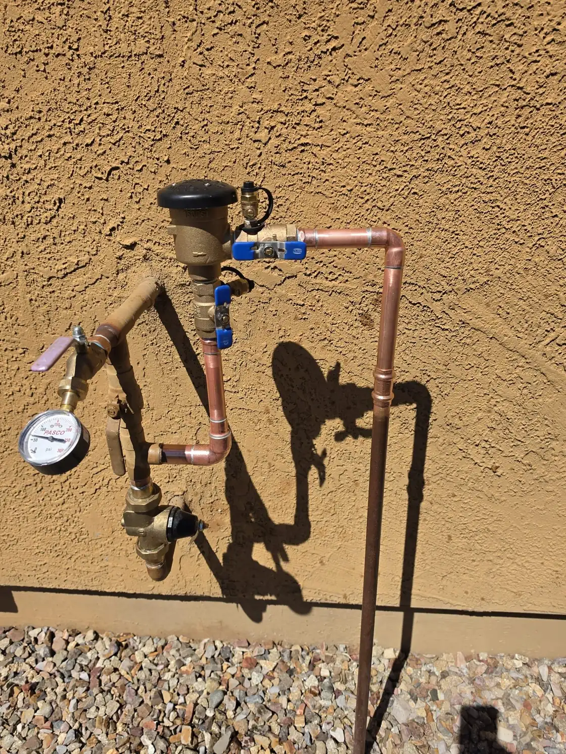 A copper supply assembly with a pressure gauge and backflow device on a tan stucco wall