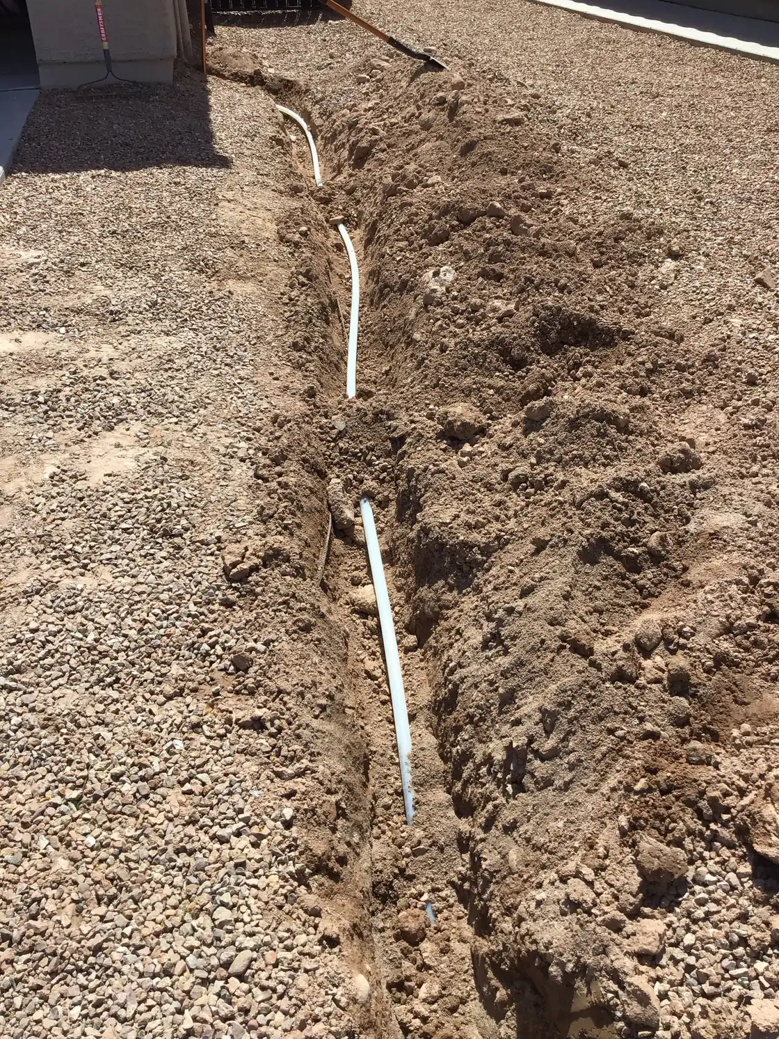 A long narrow trench through a gravel yard with a white pipe laid along it