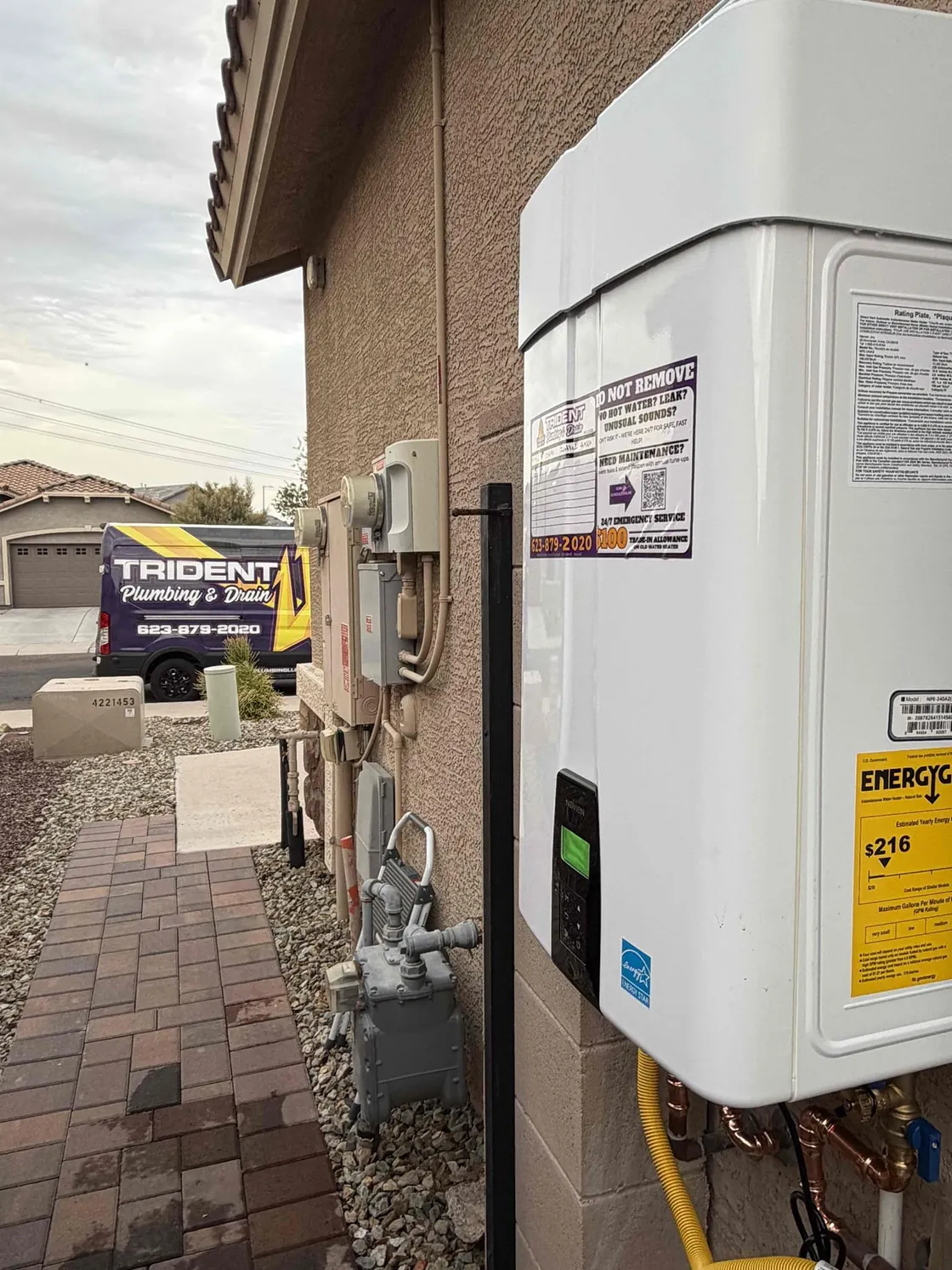 An exterior tankless water heater on a stucco wall with a Trident van parked beyond the walkway