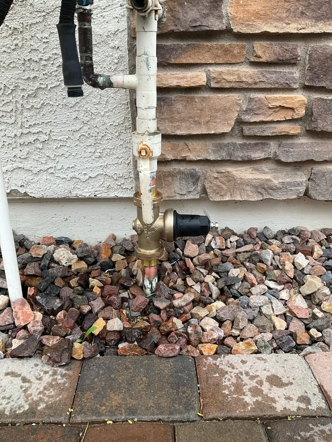 A brass pressure-reducing valve on a home's main water line beside a stone-veneer wall and a gravel bed