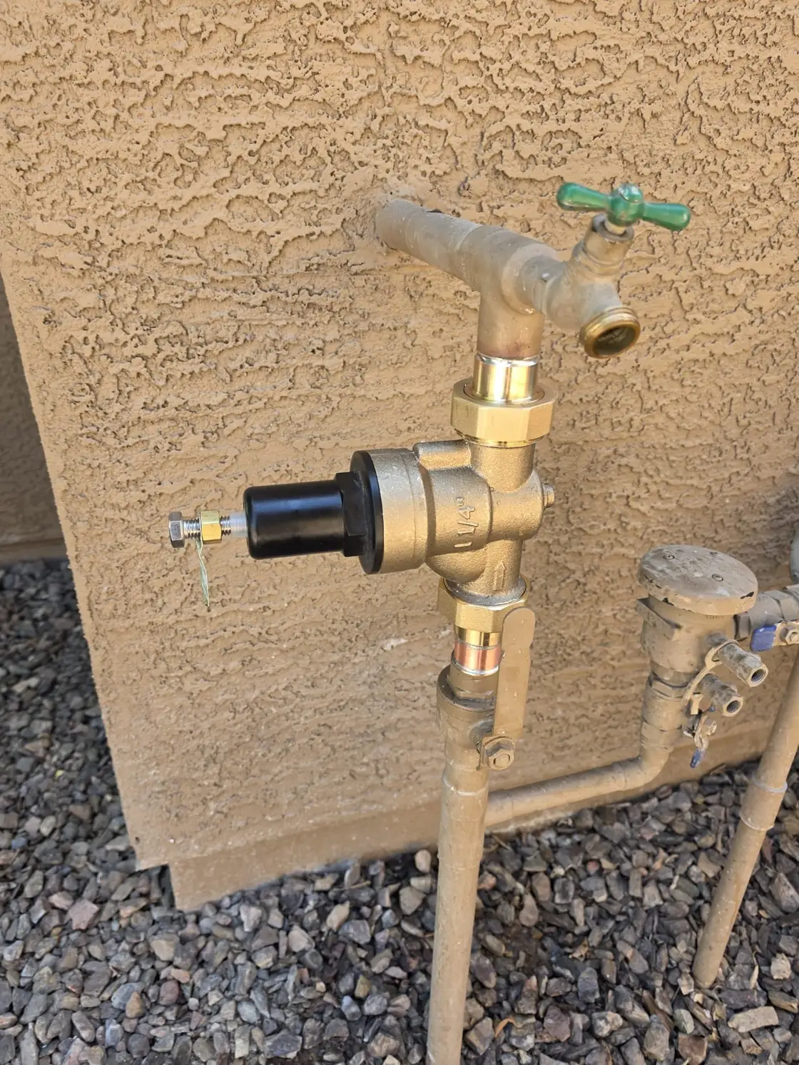 A brass pressure regulator and green-handled valve on a supply line beside a stucco wall