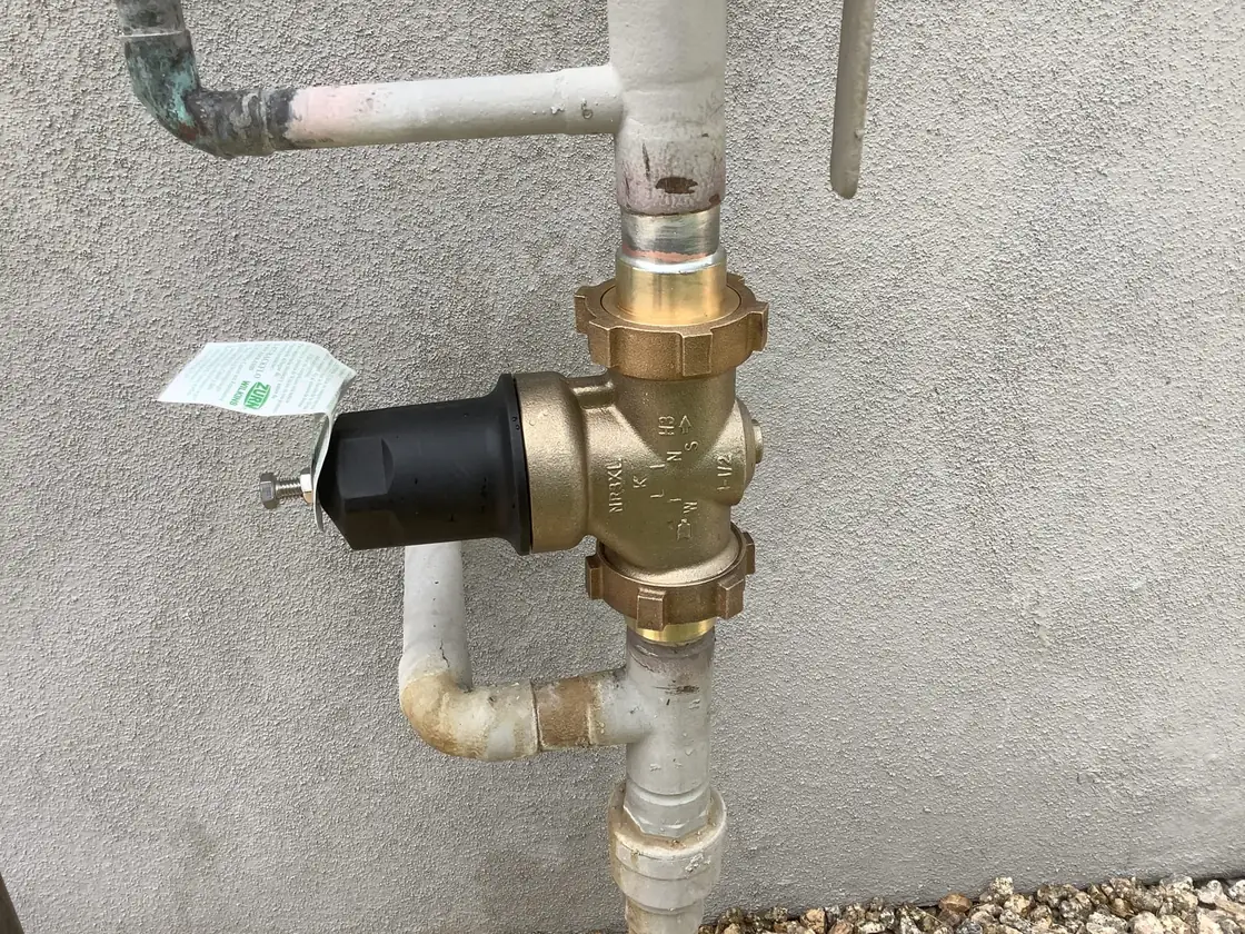 A new brass pressure regulator with its tag attached on a supply line against a gray wall