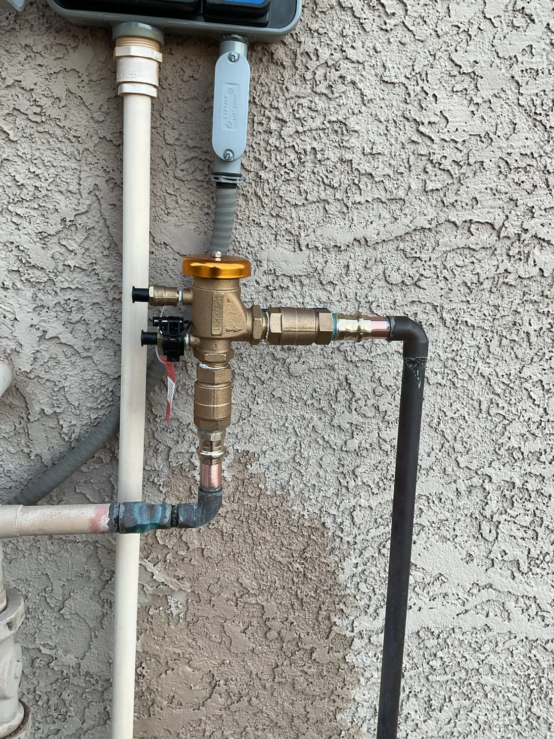 A pressure regulator assembly on a stucco wall, with a darker patch of stucco behind the piping