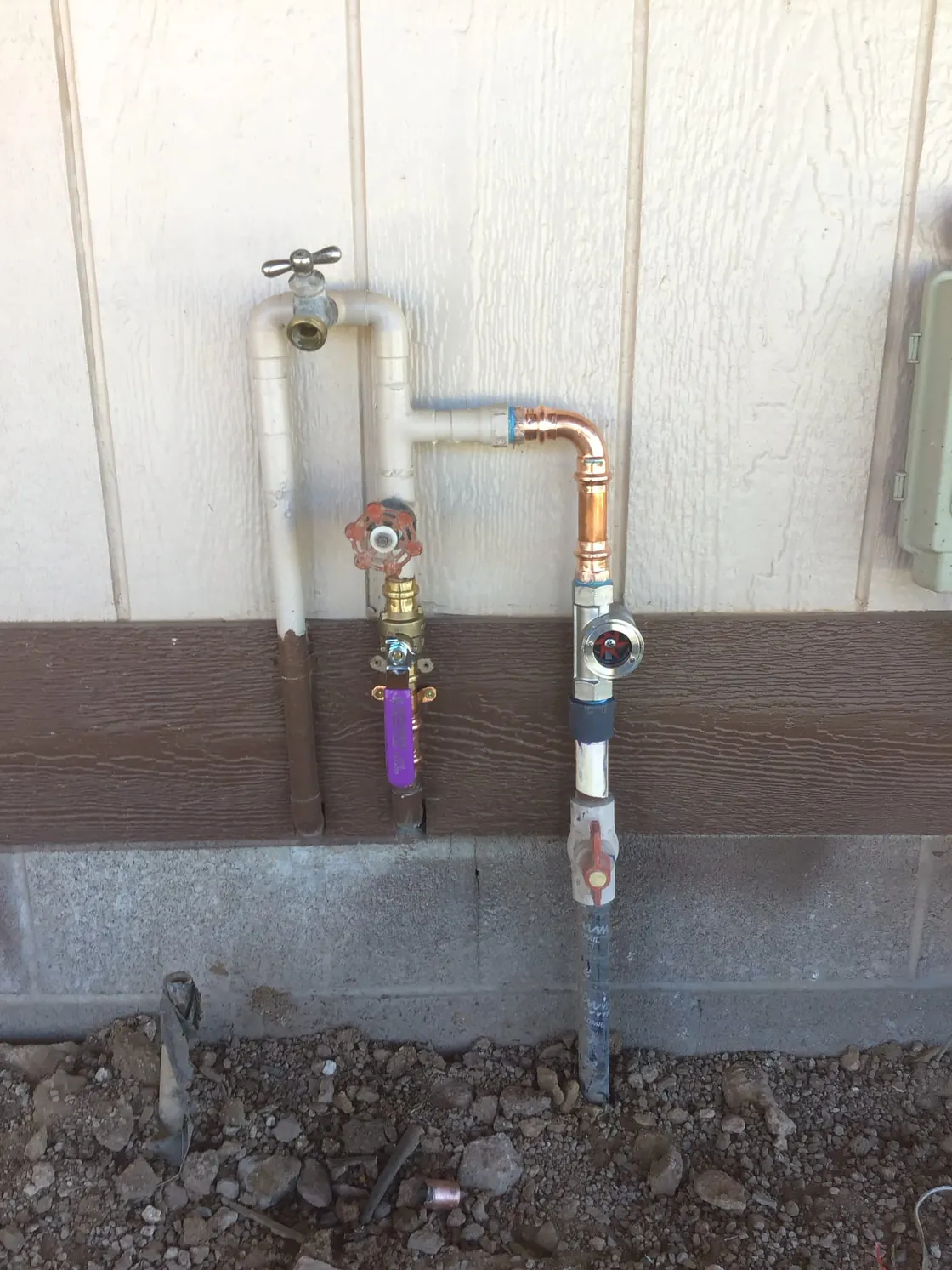 A main water supply line with a purple-handled shutoff valve and pressure regulator on a sided exterior wall