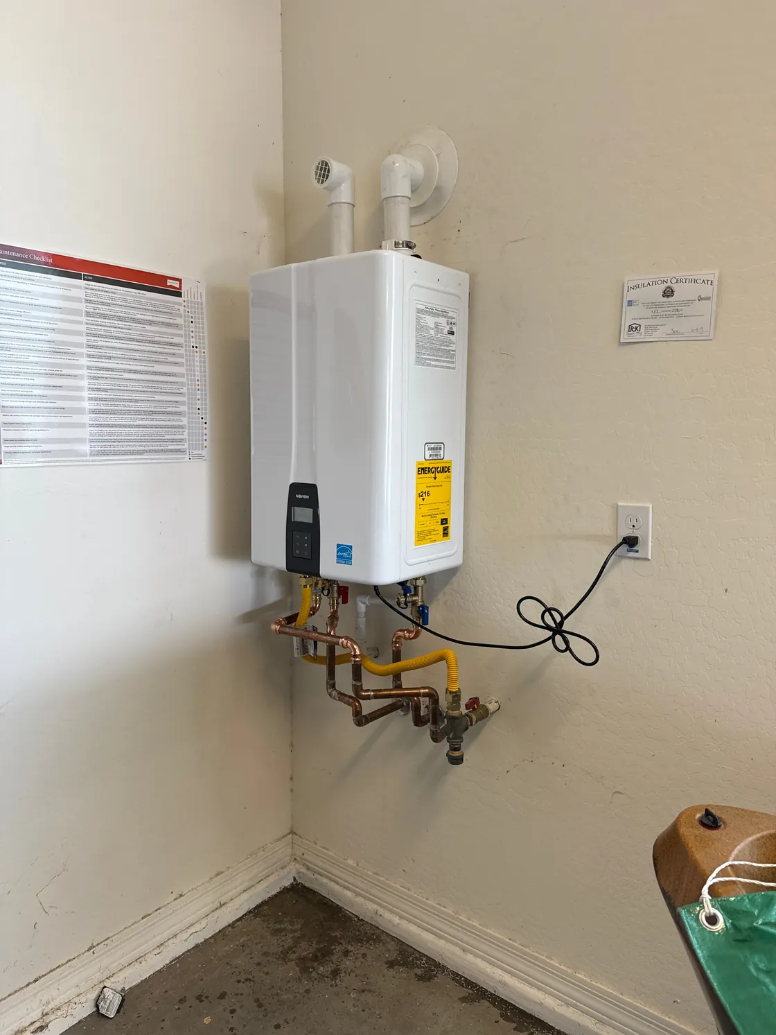 A tankless water heater in the corner of a laundry space with its connections below