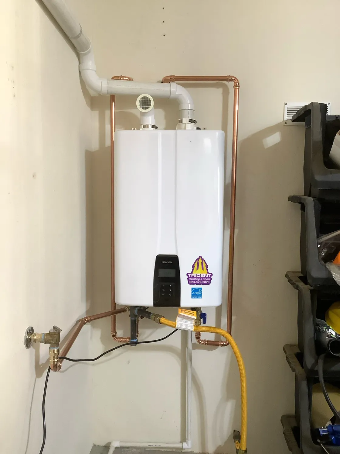 A tankless water heater with copper piping on a garage wall beside storage racks