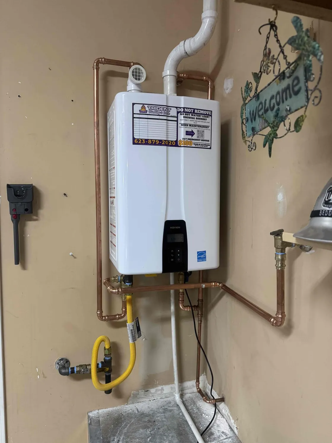 A tankless water heater on a garage wall beside a decorative welcome sign