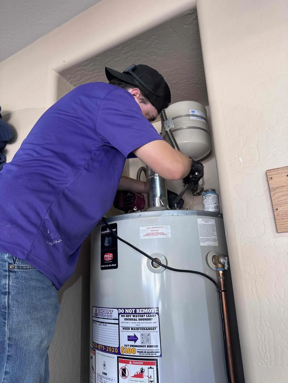 A Trident technician working on the top connections of a tank water heater in a closet