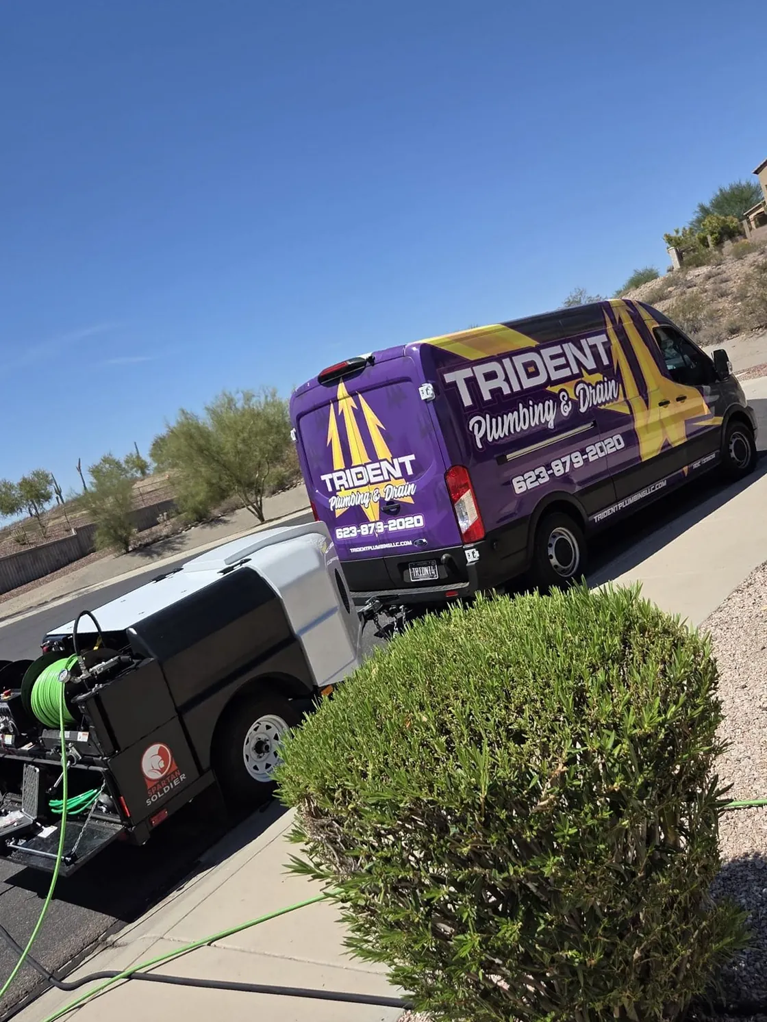 A purple and gold Trident service van and a hydro-jetting trailer parked on a sloped desert residential street