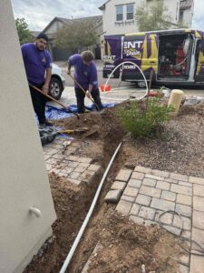 A plumber excavating and inspecting a main water line in a trench next to a brick walkway.