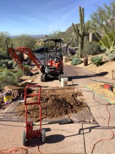 A mini-excavator digging a trench to access an underground main water line next to a brick walkway and a desert landscape.