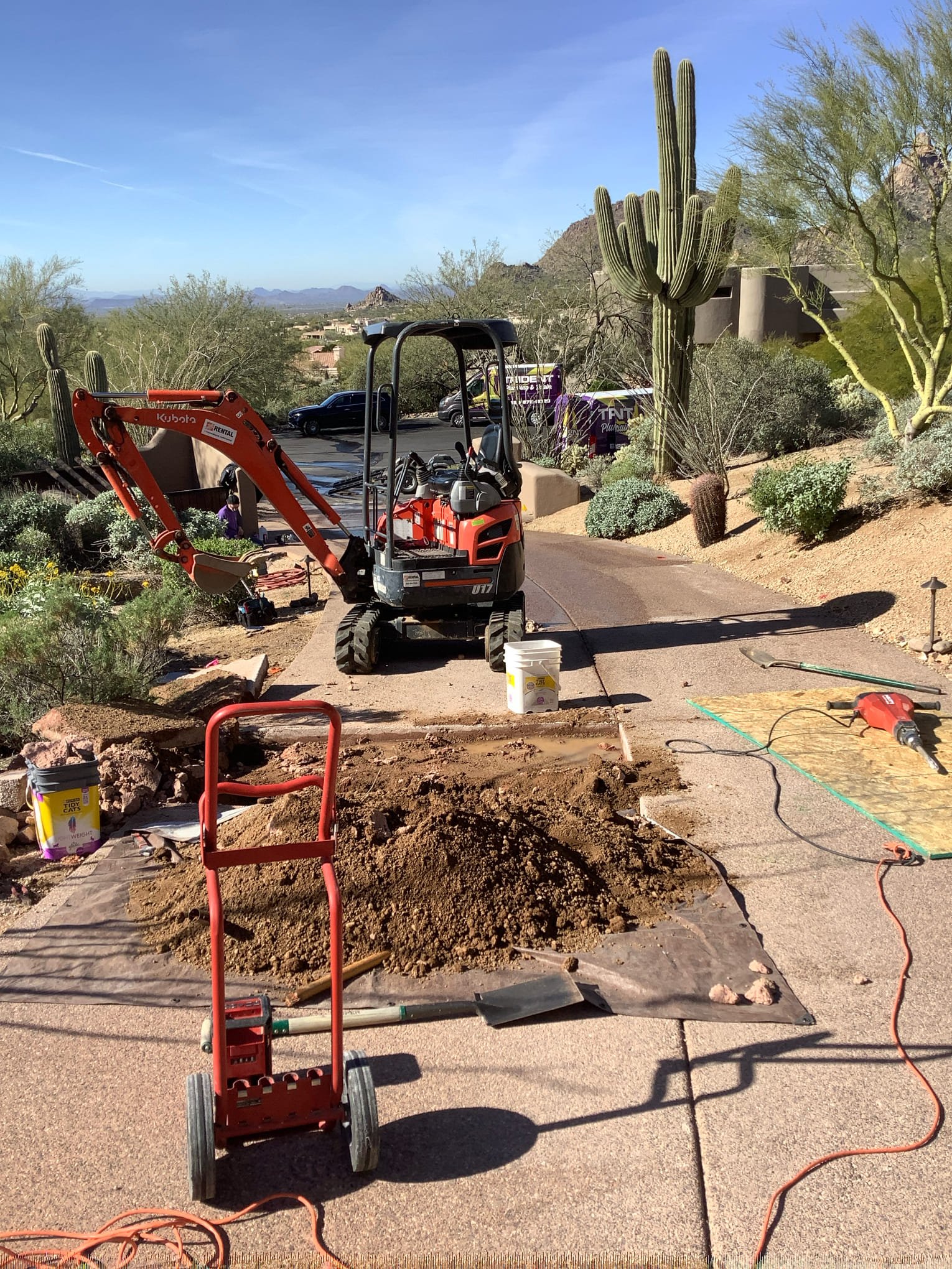 A mini-excavator digging a trench to access an underground main water line next to a brick walkway and a desert landscape.