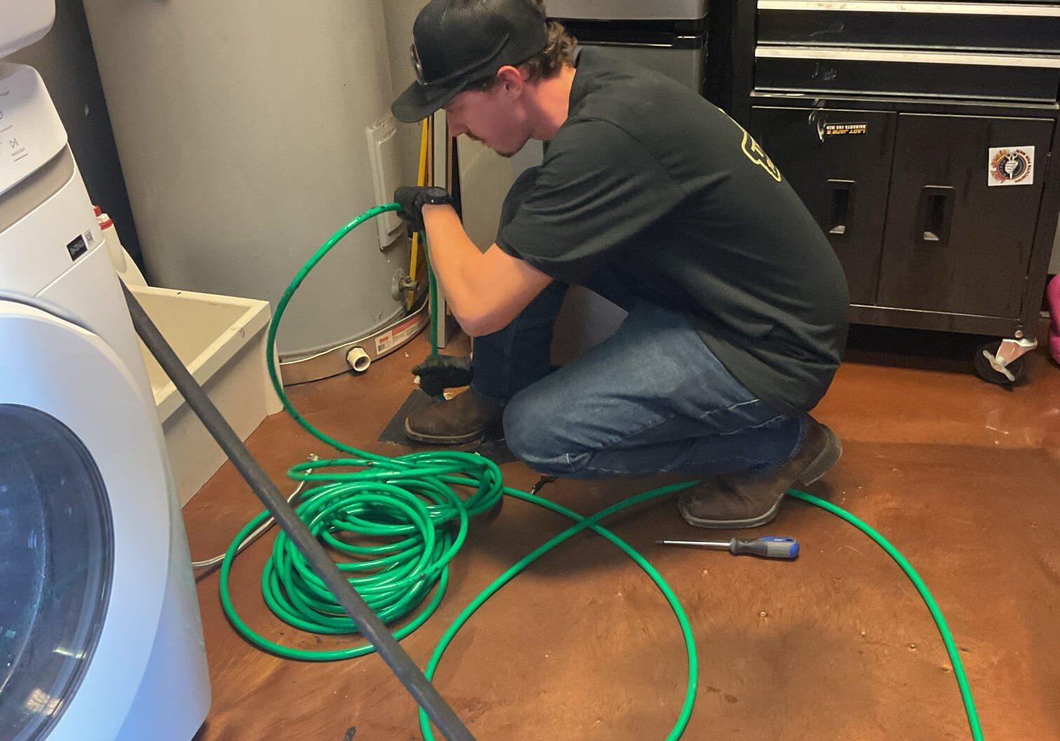 A plumber kneeling on a tile floor while carefully feeding a flexible green hydro jetting hose into a drain pipe cleanout.