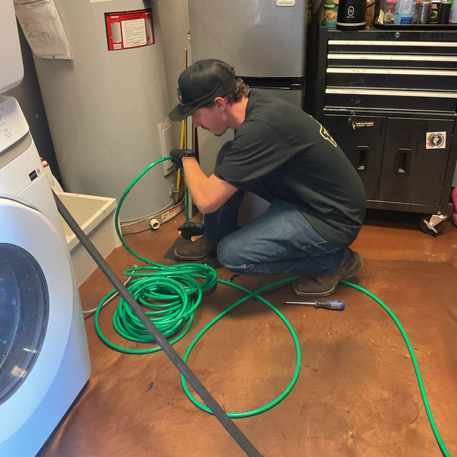 A plumber kneeling on a tile floor while carefully feeding a flexible green hydro jetting hose into a drain pipe cleanout.