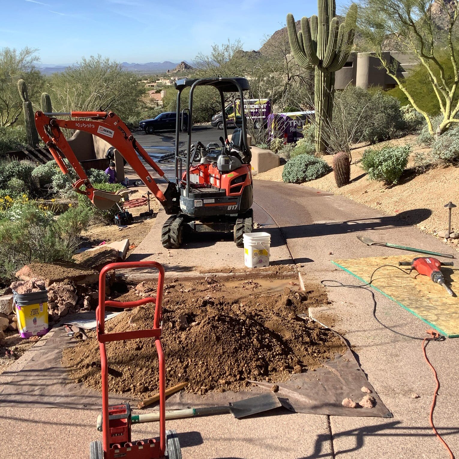 A mini-excavator digging a trench to access an underground main water line next to a brick walkway and a desert landscape.