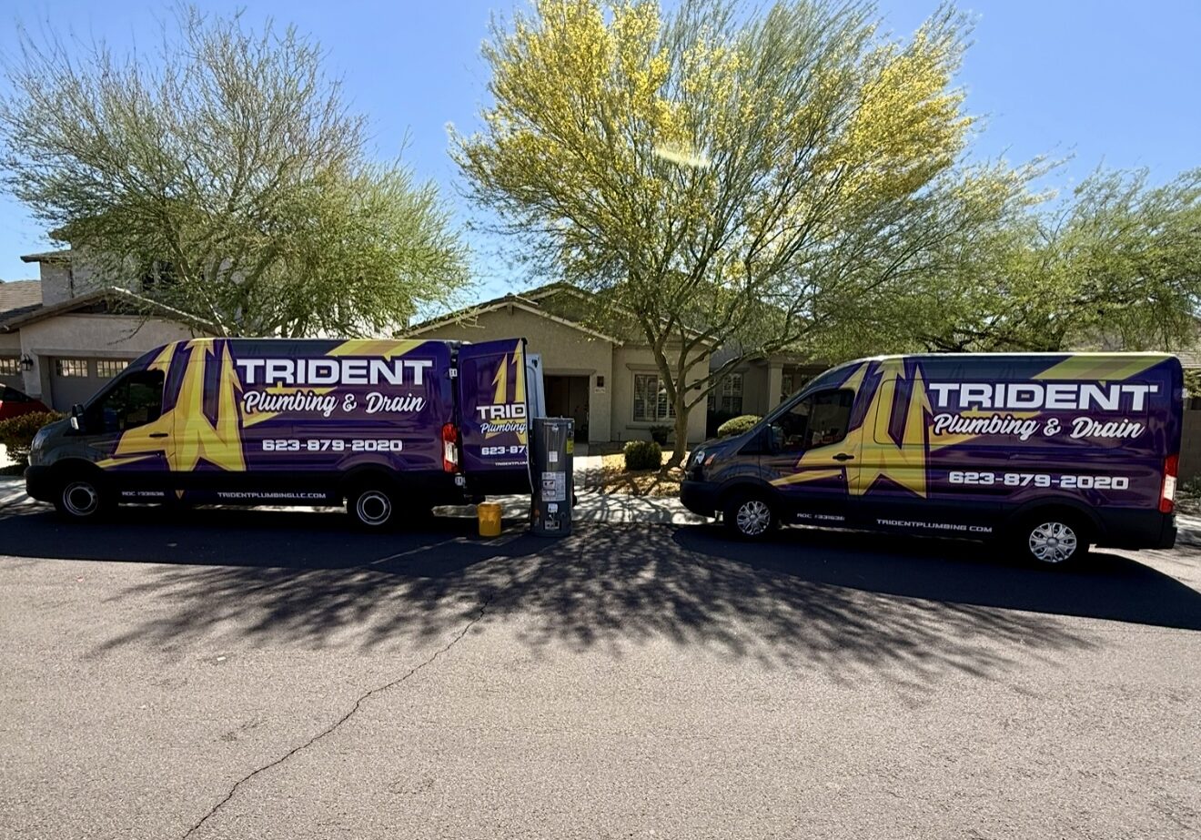 A line of yellow Trident Plumbing service vans parked in a row on an asphalt lot in Phoenix under a clear blue sky.